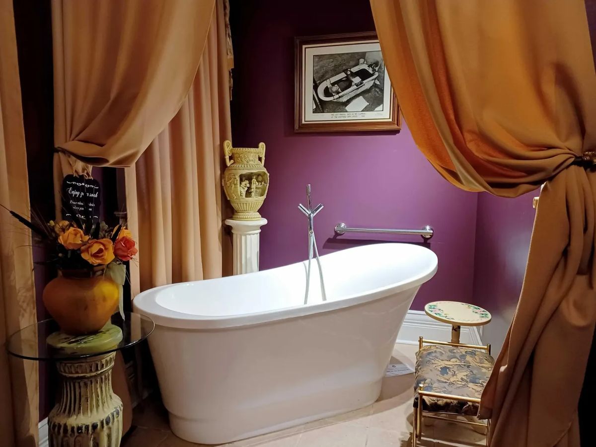 Olympia's mineral bath room: a freestanding white tub framed by gold drapes against a plum wall
